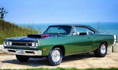 an old green car parked on the side of a road near the ocean and grass