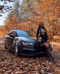 a woman sitting on the hood of a black car in front of autumn leaves and trees