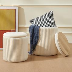 two white stools sitting on top of a wooden floor next to a painting and pillows