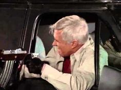 an older man sitting in the driver's seat of a car with his hand on the steering wheel