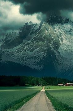 a dirt road in front of a snow covered mountain with green grass on both sides