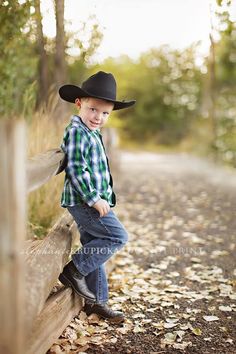 a young boy wearing a cowboy hat leaning on a wooden fence with leaves all over the ground