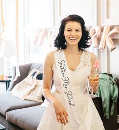 a woman in a white dress holding a wine glass and wearing a sash that says,