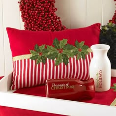 a red and white christmas pillow with holly on it, next to a bottle of body lotion