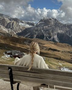 a woman sitting on top of a wooden bench in front of mountains and snow capped peaks