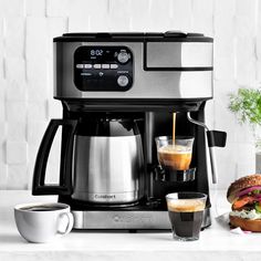 a coffee maker with two cups next to it and a sandwich on the counter top