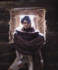 a woman standing in front of a window with a large scarf around her neck and hands on her hips