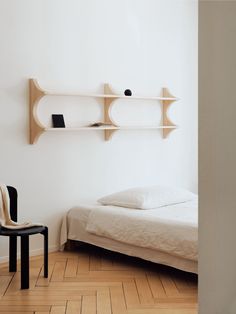 a bed sitting next to a white wall with shelves on the wall and a chair in front of it