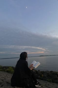 a woman sitting on the beach reading a book while looking at the sky and water