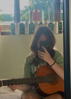 a woman sitting on a bed holding a guitar and looking at her cell phone while she is playing the guitar