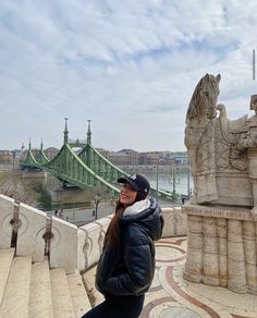 a woman in black jacket and hat standing on steps next to stone statues with bridge in background