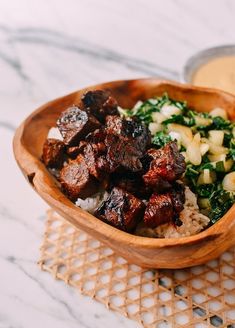 a wooden bowl filled with meat, rice and greens next to a small container of sauce