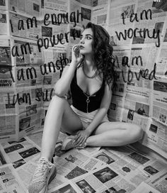 a woman sitting on the floor in front of a wall full of newspaper pages and talking on a cell phone