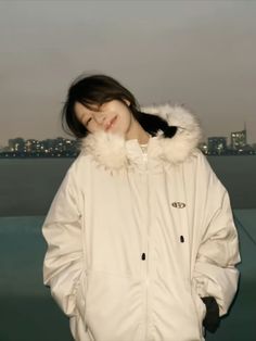 a woman standing in front of a body of water wearing a white jacket and fur collar