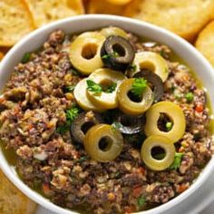 a white bowl filled with olives and ground beef dip surrounded by crackers on the side