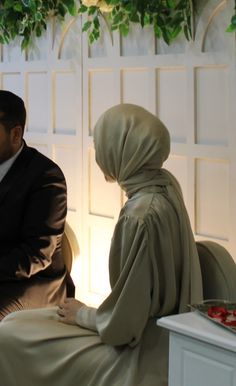 a man and woman sitting next to each other in front of a wall with plants