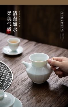 a person holding a white tea pot on top of a wooden table next to cups and saucers