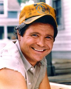 a smiling man wearing a yellow hat and white shirt sitting in front of a building