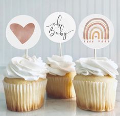 three cupcakes with white frosting and rainbow toppers, one is for a baby