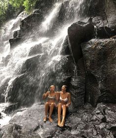 two women sitting on rocks in front of a waterfall