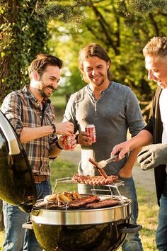 three men grilling hot dogs and drinking beverages on an outdoor bbq with trees in the background