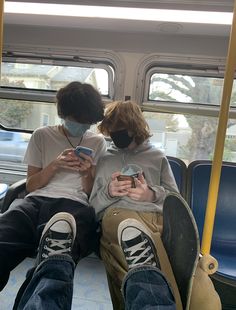 two people sitting on a bus looking at their cell phones while wearing facemasks