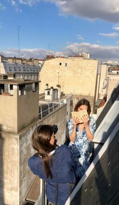 two women standing on top of a roof looking at something in front of their faces