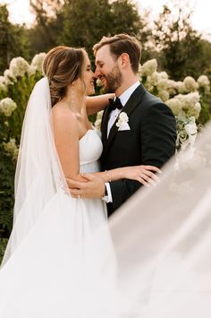 a bride and groom standing in front of white flowers with veil blowing in the wind