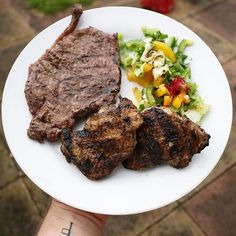 a white plate topped with meat and salad next to a person's hand holding it