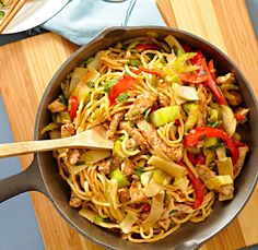 a skillet filled with noodles, meat and veggies on top of a wooden cutting board