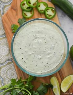 a wooden cutting board topped with a glass bowl filled with white sauce surrounded by sliced green peppers and cilantro