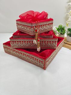 three red and gold jewelry boxes sitting on top of a white cloth covered table next to a potted plant