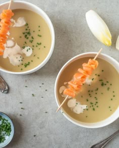 two white bowls filled with soup and vegetables on top of a gray surface next to spoons