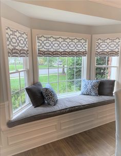 a window seat with pillows on it in front of two windows that have roman shades