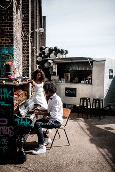 two people sitting at a piano in front of a building with graffiti on the walls