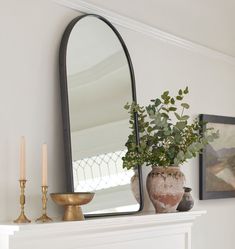 a white mantle with a mirror, vase and candles on it next to a potted plant