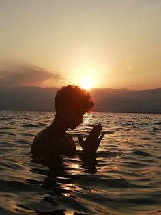 a man sitting in the water with his hands together as the sun sets behind him