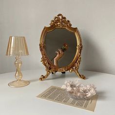 a mirror and lamp on a table with a hand holding a flower in front of it