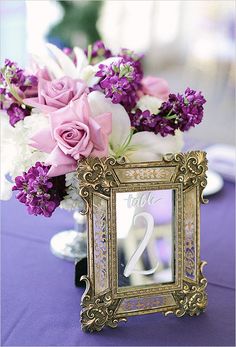 purple and white flowers are in a gold frame on a table with a blue cloth