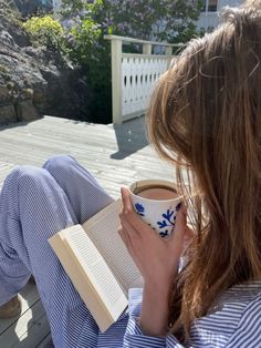 a woman sitting on a deck reading a book and holding a coffee cup with an open book in her lap