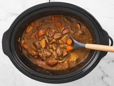 a crock pot filled with beef stew and carrots next to a wooden spoon