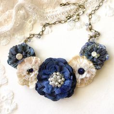 Classic colors blue and white are always in fashion. This eye-catching fabric flower bib necklace features layers of hand-cut and sewn textiles in blue, ivory and cream white hues. Pretty textures and mixed patterns, along with vintage lace, unique antique glass buttons and jewels make this a one-of-a-kind accessory. Pretty Czech glass beads adorn the oval link chain. Adjustable to any length. * Flower ~ 7 x 2.5 inches * Length ~ 26 inches long, adjustable with locking clasp. An extender chain c Floral Necklace Jewelry, Rosy Posy, Fabric Flower Necklace, Necklace Fabric, Fabric Flower Pins, Floral Statement Necklace, Mixed Patterns, Textile Necklace, Bride Necklace