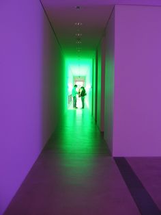 two people walking down a long hallway with green and purple lights on the walls in front of them