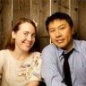 a man and woman sitting next to each other in front of a wooden wall with strings hanging from it