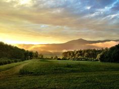 the sun is setting over some trees and hills in the distance, with clouds hovering overhead