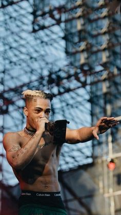 a shirtless man with tattoos on his chest and hands in the air, holding something up to his mouth