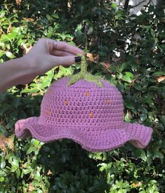 a hand holding a pink crocheted hat with oranges on it in front of some bushes