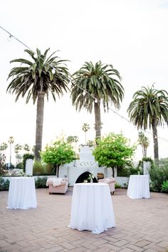 tables with white tablecloths are set up in front of palm trees and an outdoor fireplace