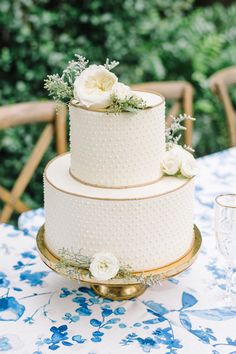 a three tiered cake with white flowers and greenery sits on a gold platter