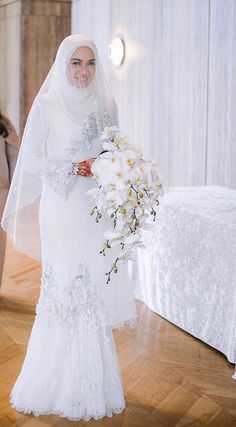 a woman in a white wedding dress holding a bouquet of flowers and standing next to a bed